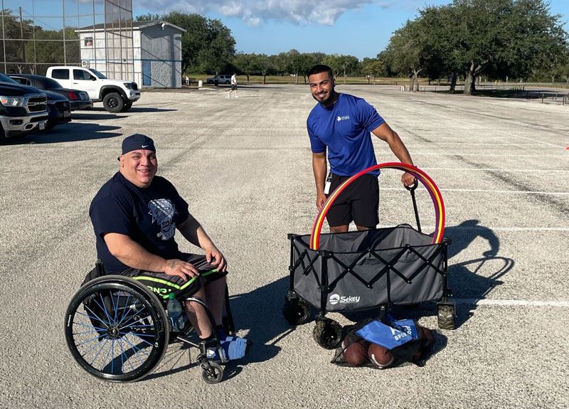 Two men in a parking lot, one in a wheelchair one standing, with a wagon of hula hoops and footballs