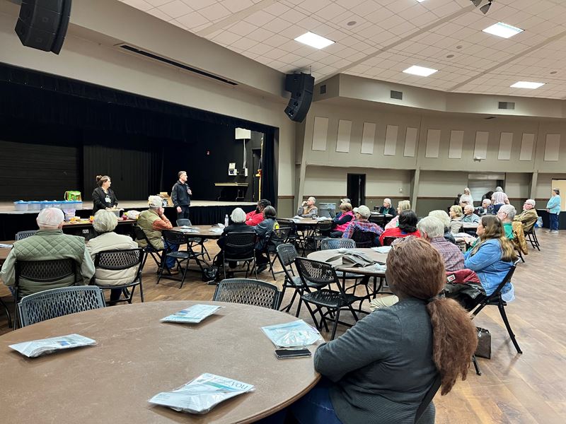 Several people sit at round tables at the community center dome watching two speakers