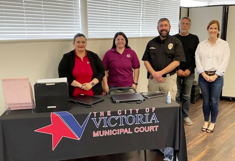 Five people stand behind a table with a City of Victoria Municipal Court tablecloth and laptops.