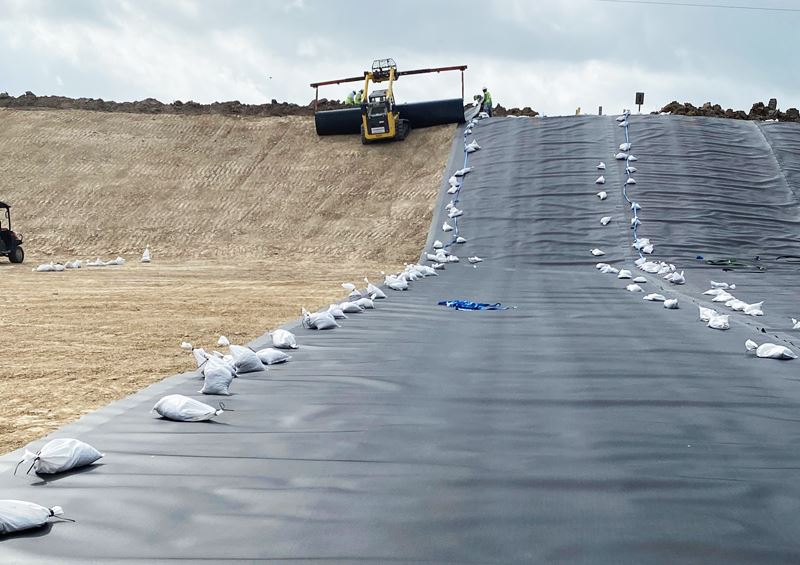 A machine rolls out a black plastic covering over a smooth dirt surface.
