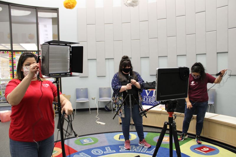 Three women wearing face masks setting up video equipment in a room with a colorful carpet
