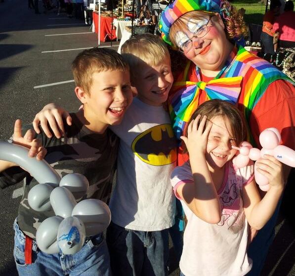 A clown wearing face paint and rainbow clothes poses with kids holding balloon animals