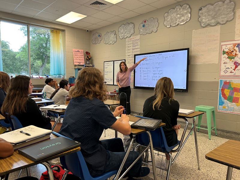 A woman in front of an eight-grade class points to a smartboard.