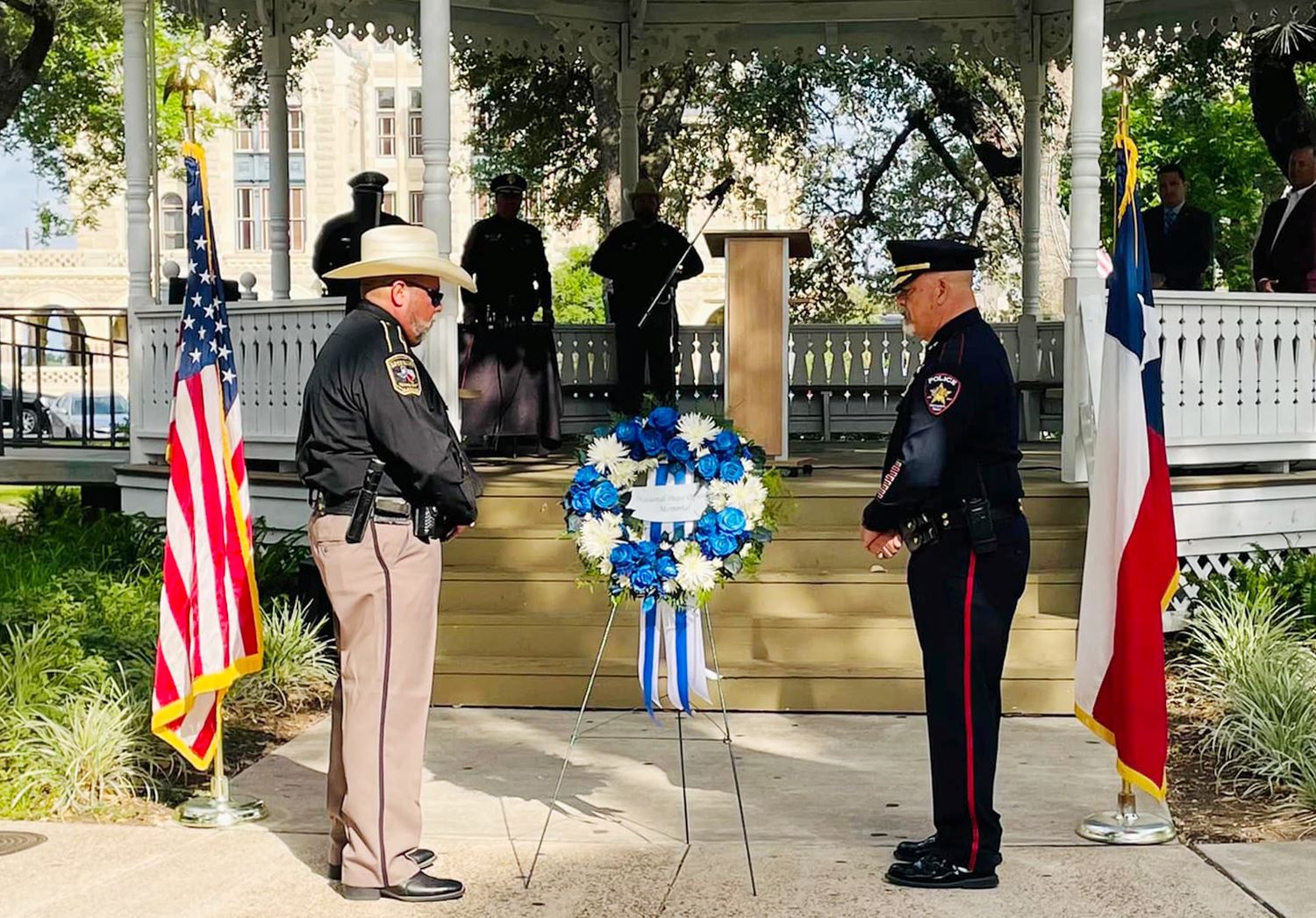 Two men in law enforcement uniform stand on either side of a blue and white wreath, heads bowed