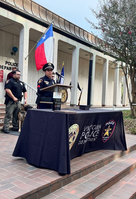 A woman in police uniform speaks at a podium in front of the Police Department building