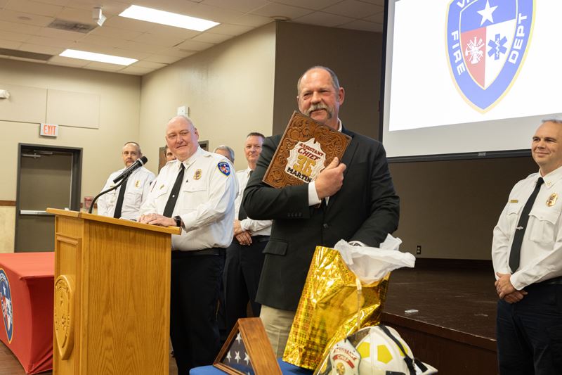 Chief Shannon Martin holds an engraved plaque or book, surrounded by firefighters in dress uniform
