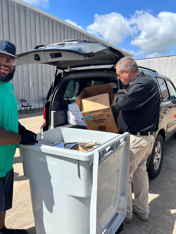 A man putting a box in the trunk of a car. Another man standing next to a trash bin.