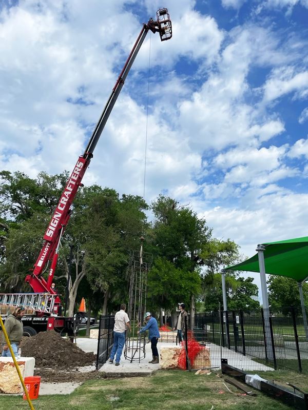 In front of the dog park, a Sign Crafters crane is used to lift a tall metal frame into place