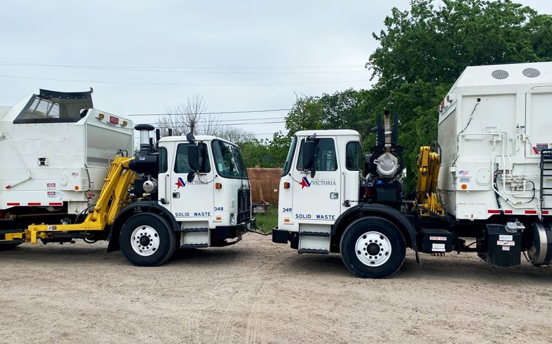 Two white trash pickup trucks with the City of Victoria logo and Solid Waste written on them