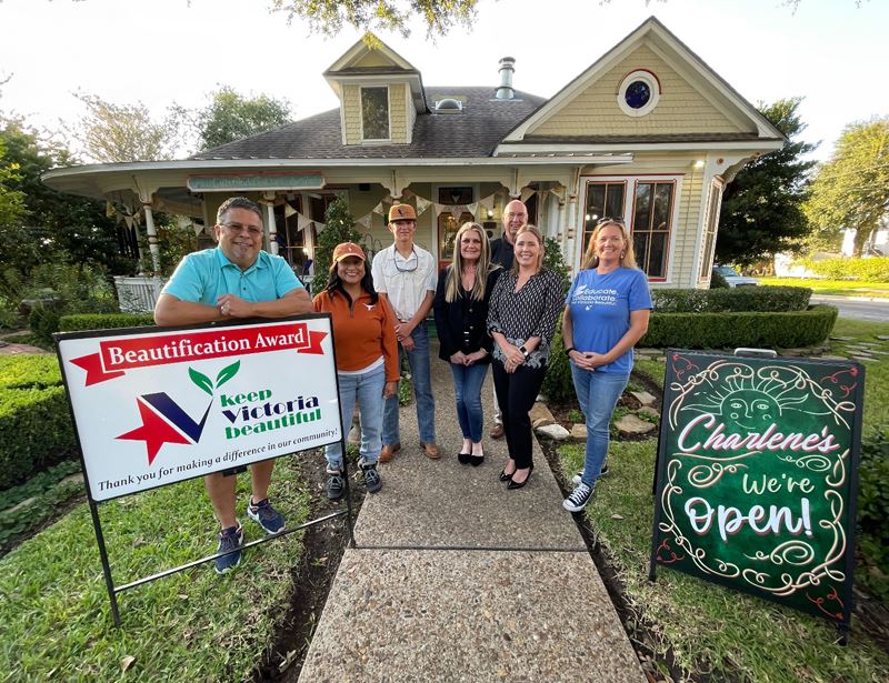 Group photo with KVB sign in front of an older style home with bright trim and neat shrubbery