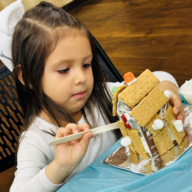 A girl decorating a small gingerbread house made of graham crackers on a milk carton