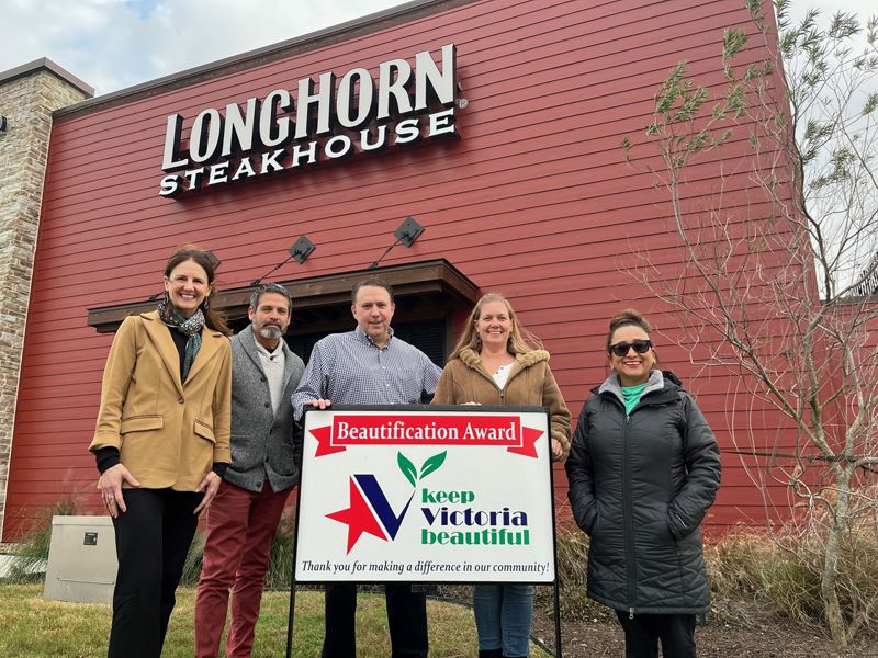 Group photo with KVB sign in front of LongHorn Steakhouse. Flowerbed with tree and shrubs