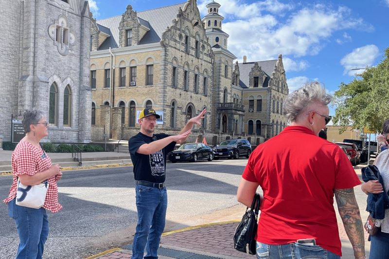 A man in a black T shirt and jeans gestures while talking to a group in front of St. Mary's downt
