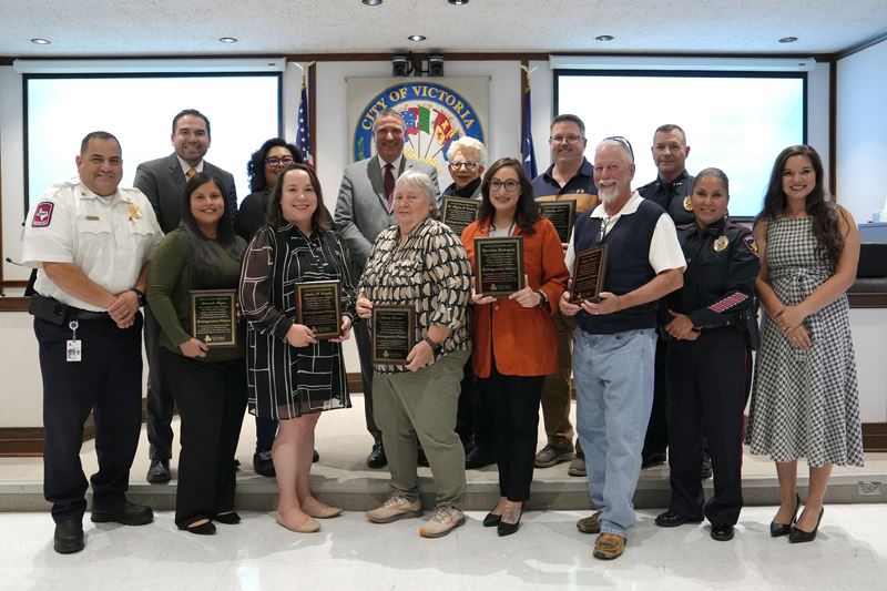 Large group photo of people holding plaques along with City officials at Council chambers.