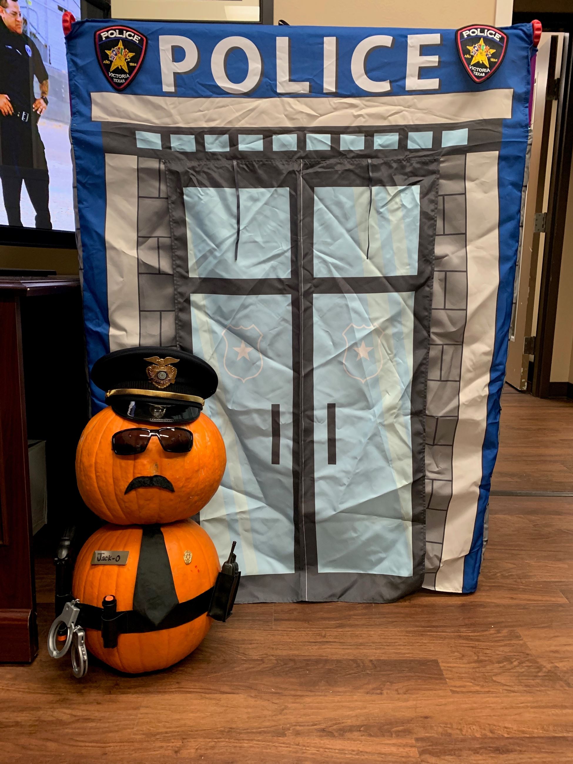 Pumpkin figure dressed as a policeman in front of a police station backdrop. Name tag reads 