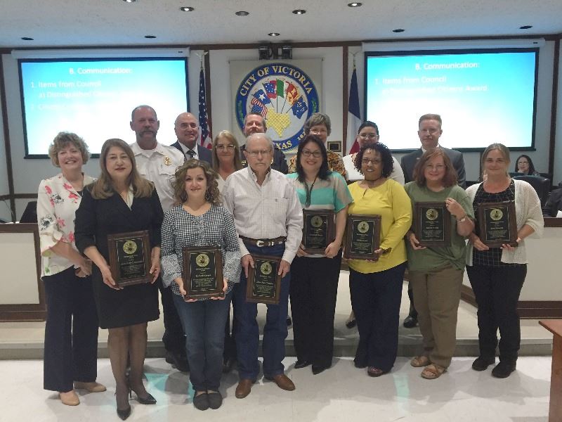 A group of adults pose with plaques in City Council chambers surrounded by mayor and other officials