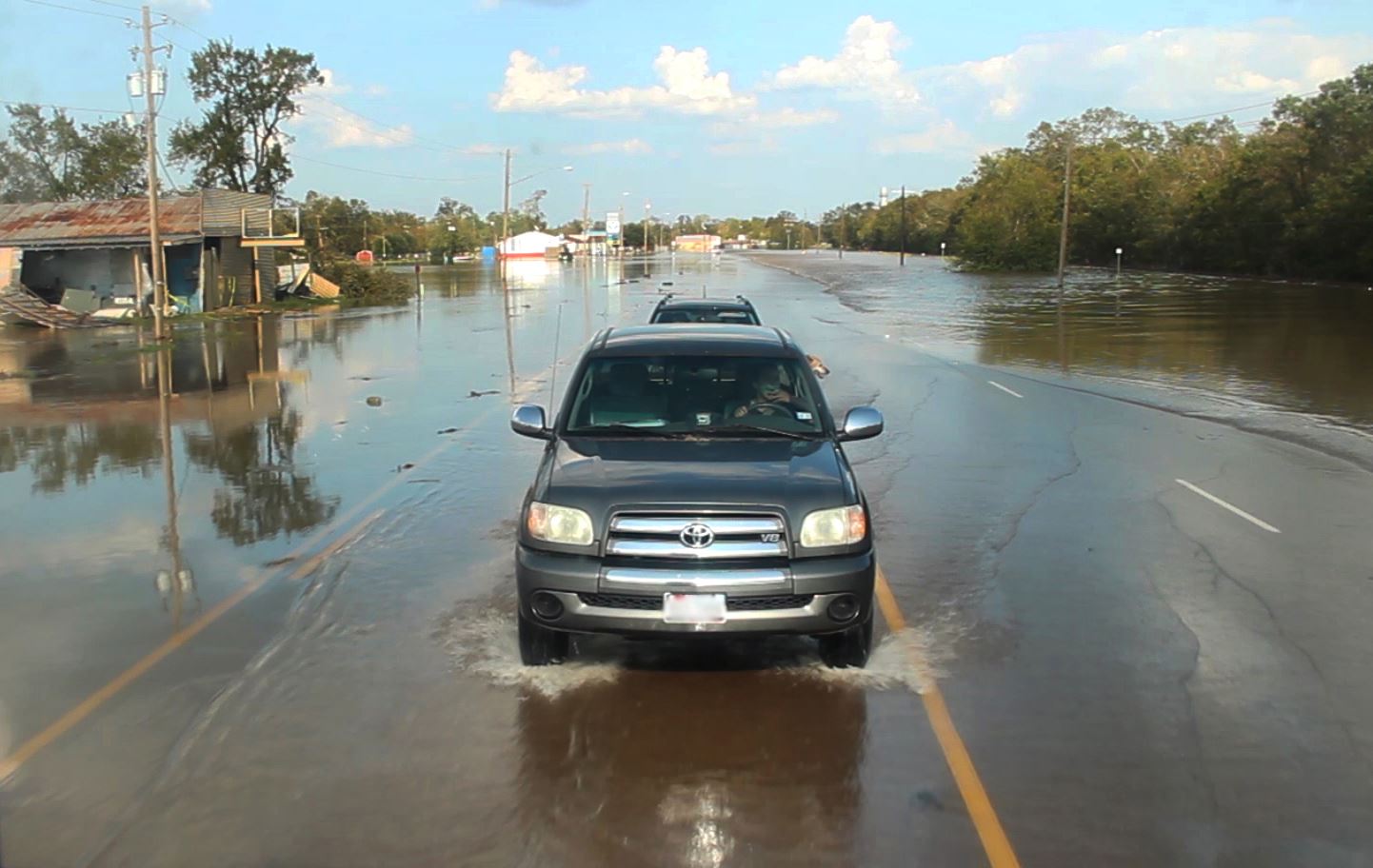 Truck drives down flooded road past flooded buildings. A dog hangs its head out the window.