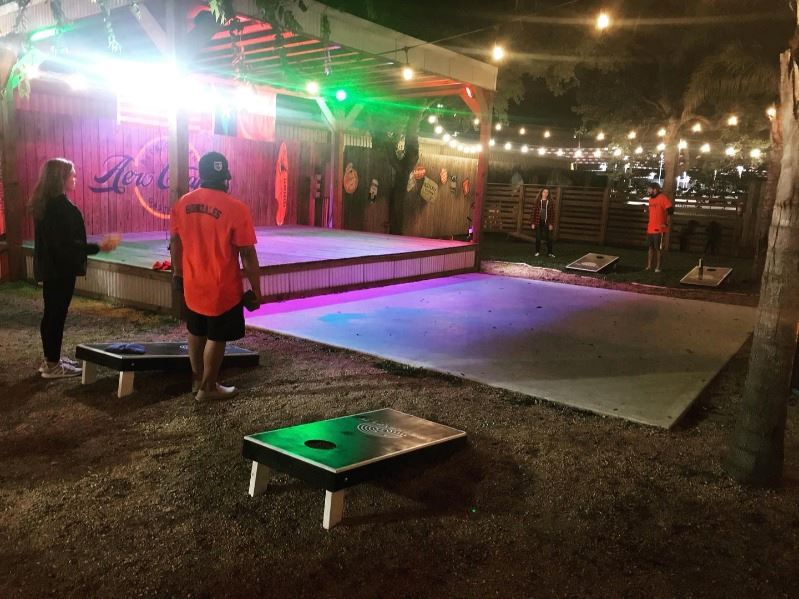 People play cornhole on an outdoor patio at night beneath string lights near Aero Crafters stage.