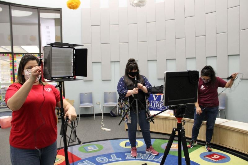 Three women set up video and lighting equipment in a room with a colorful carpet.