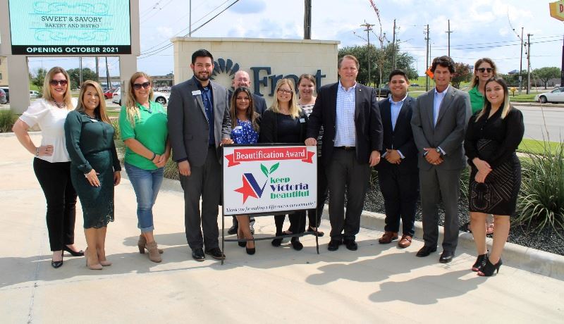 Group of people, some in business attire, pose with KVB sign near Frost Bank sign and landscaping.