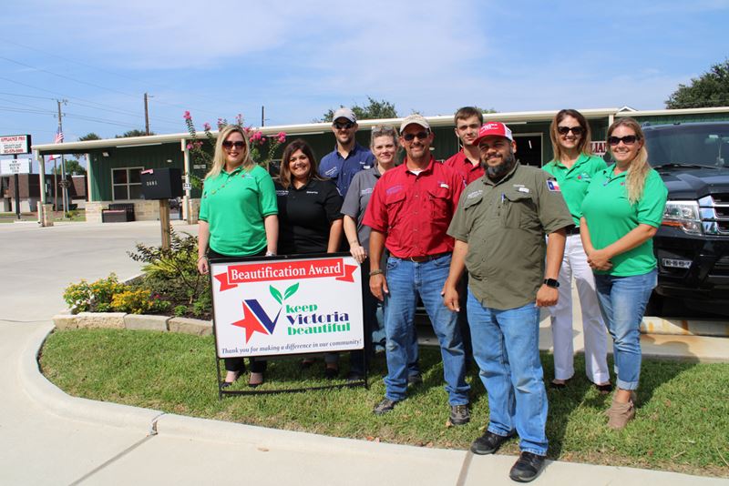 Group photo with KVB sign in front of Appliance Pro. Flowering plants and trees are visible.