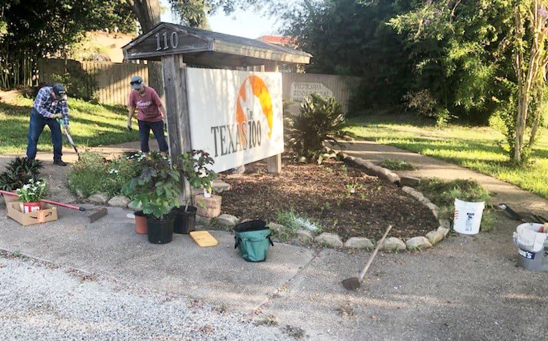 Two people work in a flowerbed at the Texas Zoo sign surrounded by potted plants