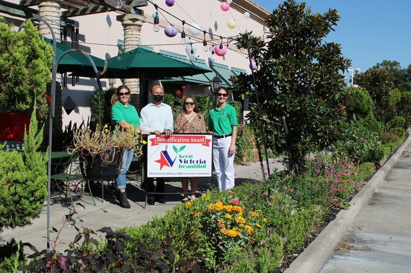 Four people stand with KVB sign on patio decorated with flowers, shrubs and paper lanterns.