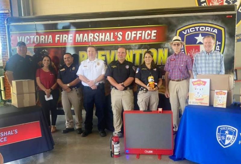 Group photo with Fire Department Fire Marshal backdrop. Sparky the Dog themed items in foreground.