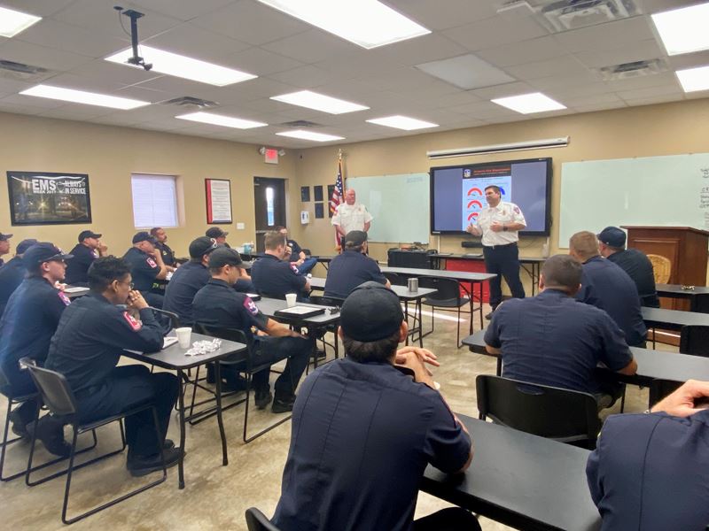 Firefighters sit at rows of tables and watch two firefighters give a slide show presentation