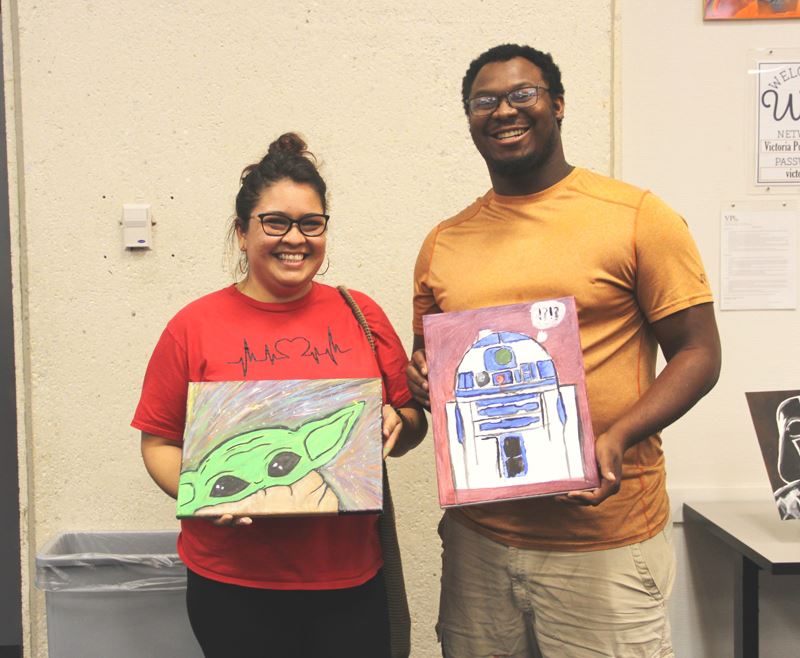 Two people hold paintings featuring Baby Yoda and R2D2 from Star Wars