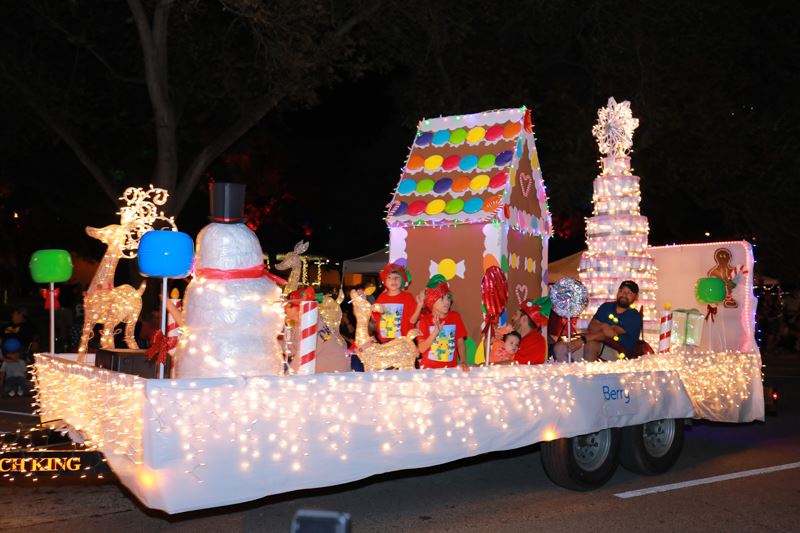 Children in elf hats on a lighted parade float with a snowman, reindeer and a gingerbread house