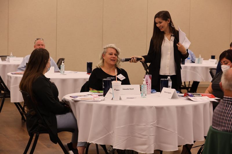 People are seated at a round table with a tablecloth. A standing woman holds mic for seated woman.