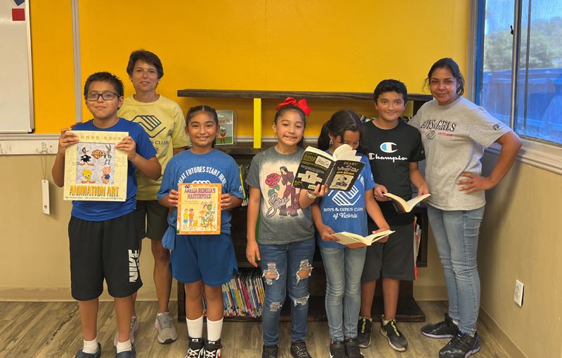 Kids and two adults pose with books in front of a bookshelf