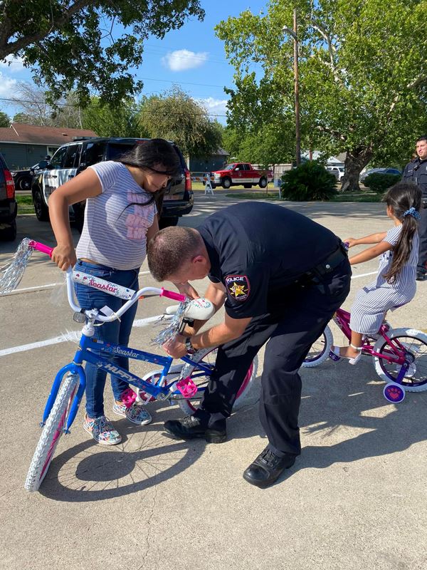 A police officer adjusts a girl's bike seat while a younger girl pedals in the background