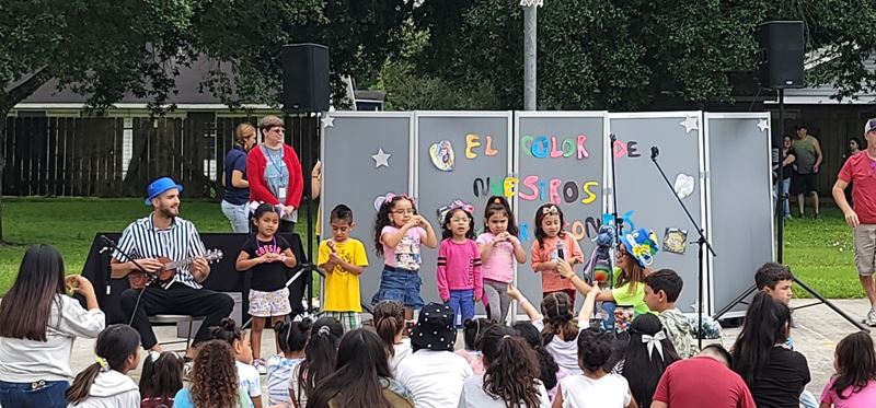 Children stand on a paved surface in a park doing a performance for seated children.