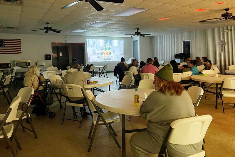 People sit at foldout tables in a large indoor dining area and watch Elf on a wall projector.