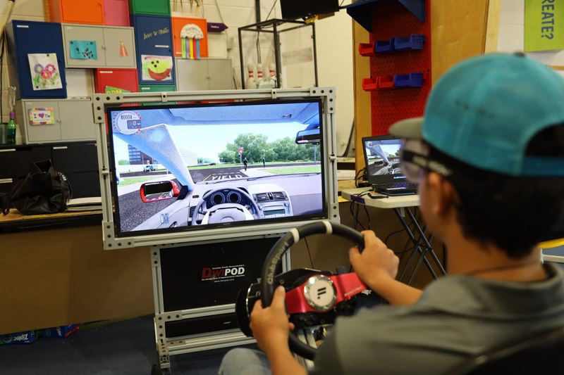 A child operates a driving simulator that includes a screen and a steering wheel controller