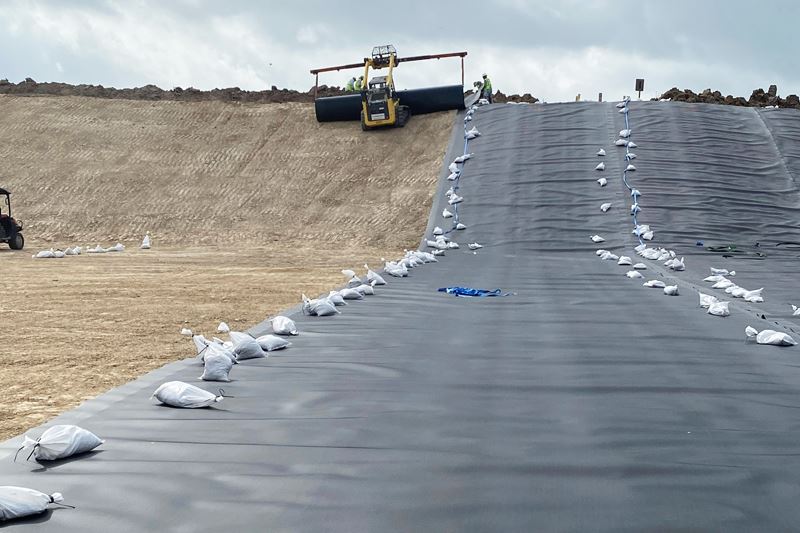 Heavy machinery is used to roll out wide, black plastic liner on a flat dirt surface.