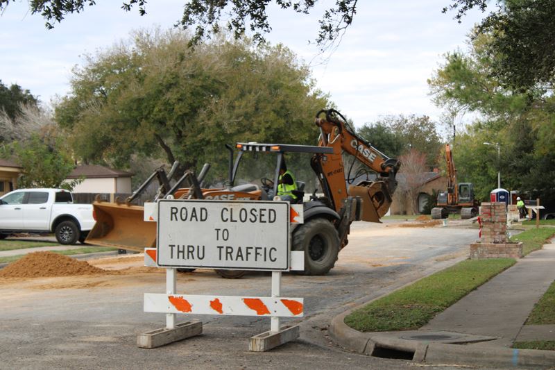 Heavy equipment operates on a barricaded road in a neighborhood. Dirt is dug up at the edges.