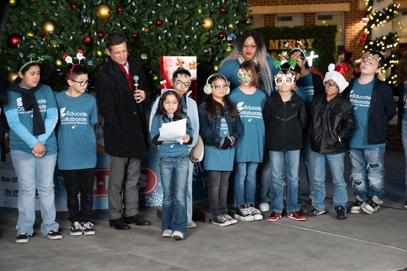 Young kids, a man and a woman stand by a giant Christmas tree. The kids and woman wear blue shirts.