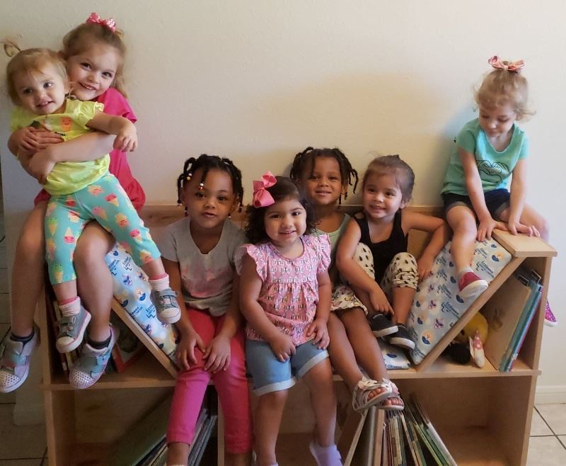 Preschool children pose with wooden reading nook that contains shelves full of books.