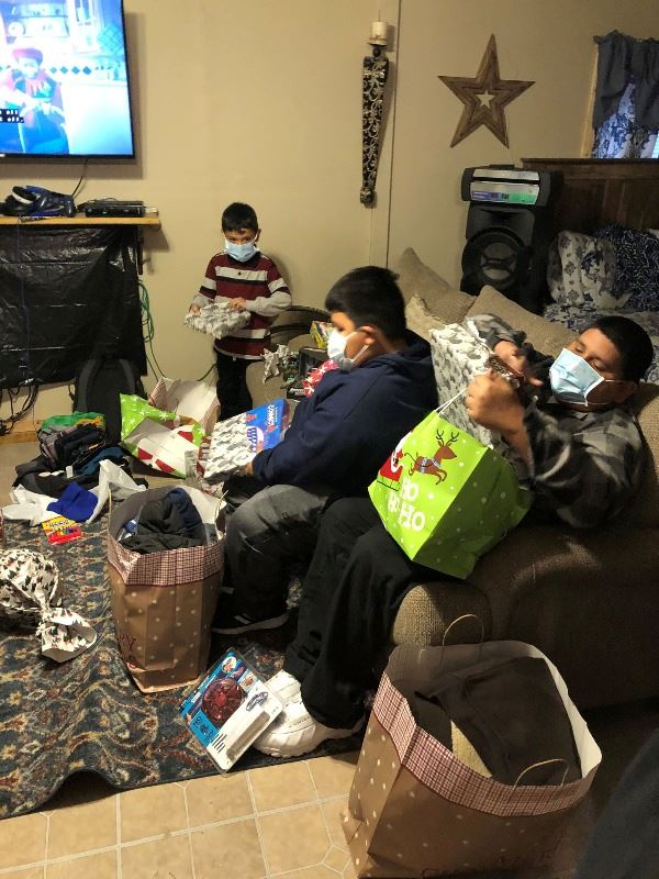 Three boys wearing face coverings open wrapped and bagged Christmas gifts in a living room.