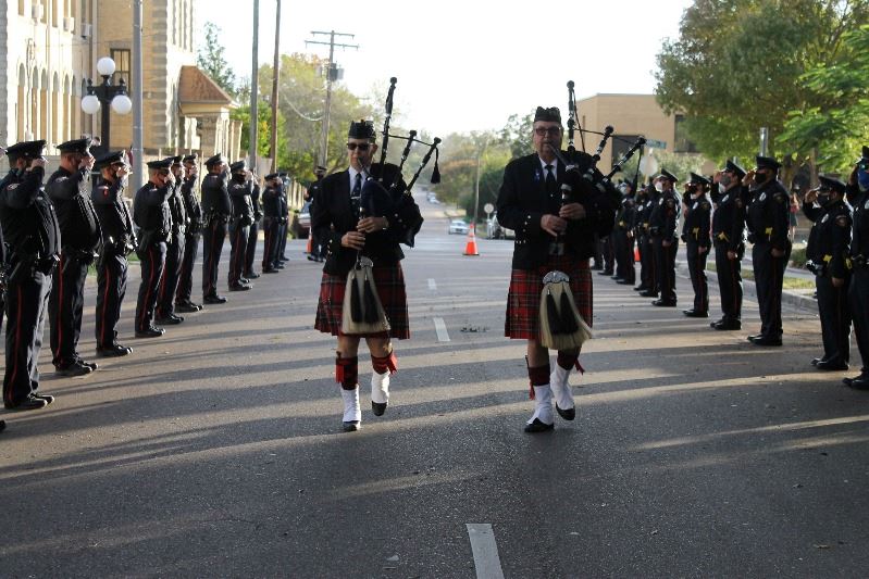 Two bagpipe performers walk down the street, accompanied by masked police officers