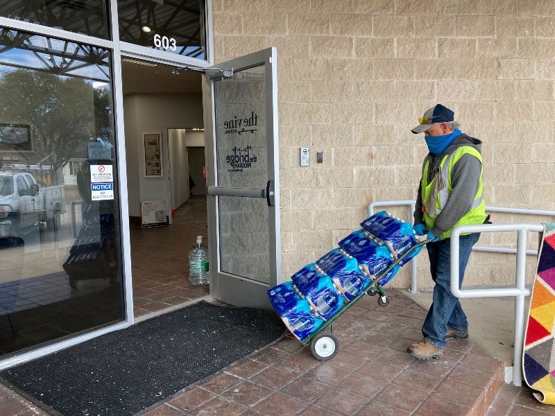 Man pushes dolly of bottled water through open glass door marked The Vine School.