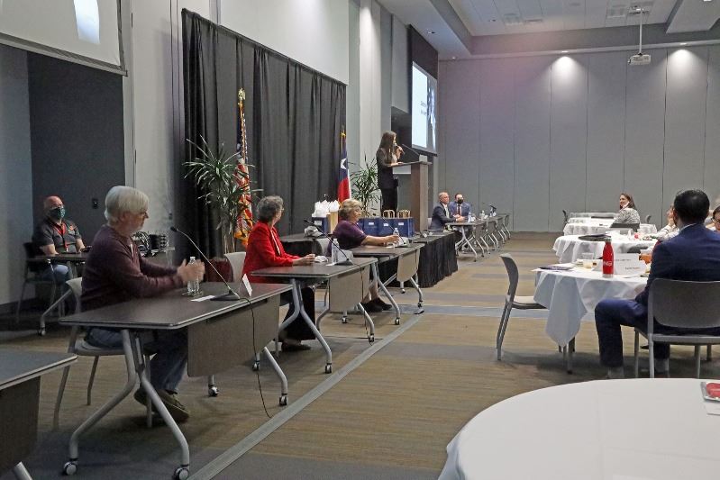 Five people sit at spaced-apart tables at the front of a ballroom. A woman stands at a podium.