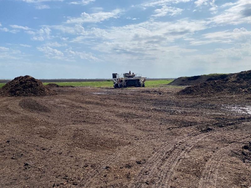 A compost turner stands in a wide, flat field of compost with a couple of hills.