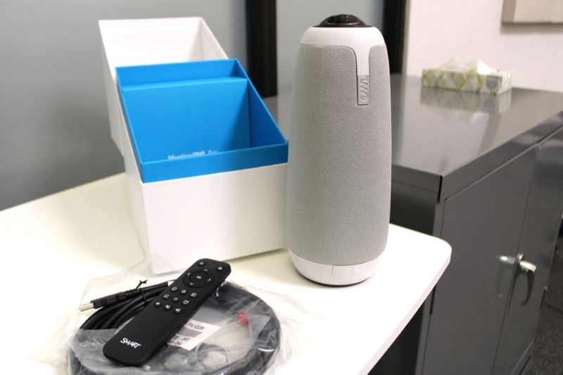 A desktop speaker/camera that resembles a smarthome speaker sits on a counter next to a remote.