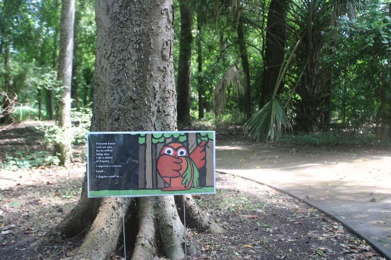 A sidewalk leads through a wooded nature trail. A printed sign shows pages from a children's book