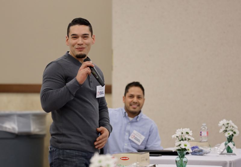 Standing man wearing name tag holds microphone and smiles.