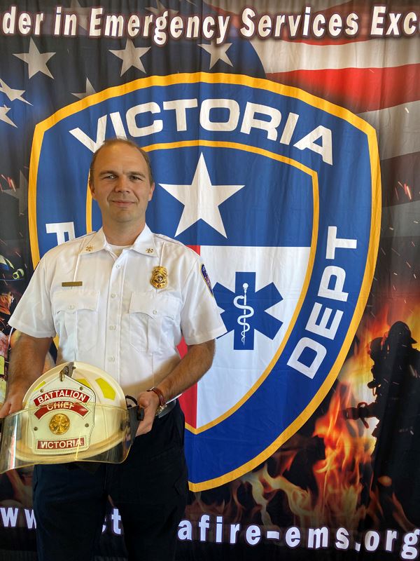 Man stands in front of fire department backdrop holding a helmet that reads Battalion Chief.